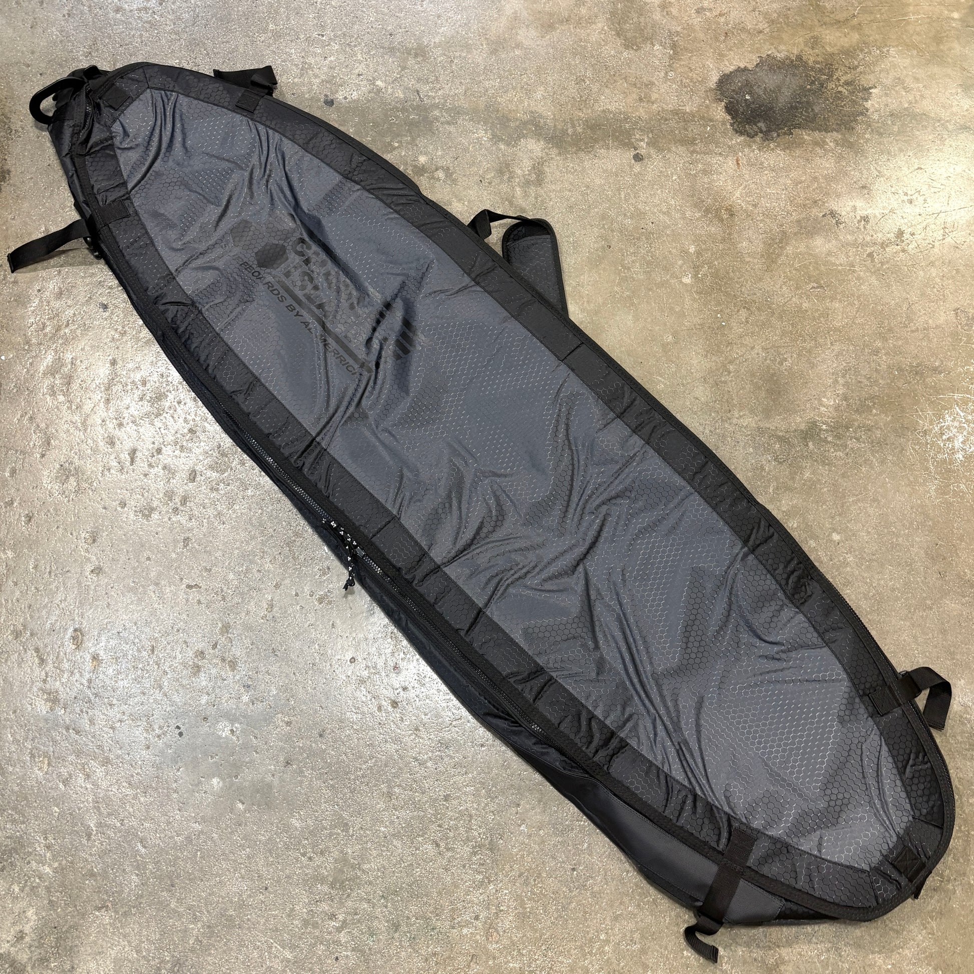 A long, narrow black surfboard bag or travel luggage cover laid diagonally on a concrete floor. The bag has a textured dark gray center panel, black trim, and black straps, with a faint gray logo printed on the upper left side.