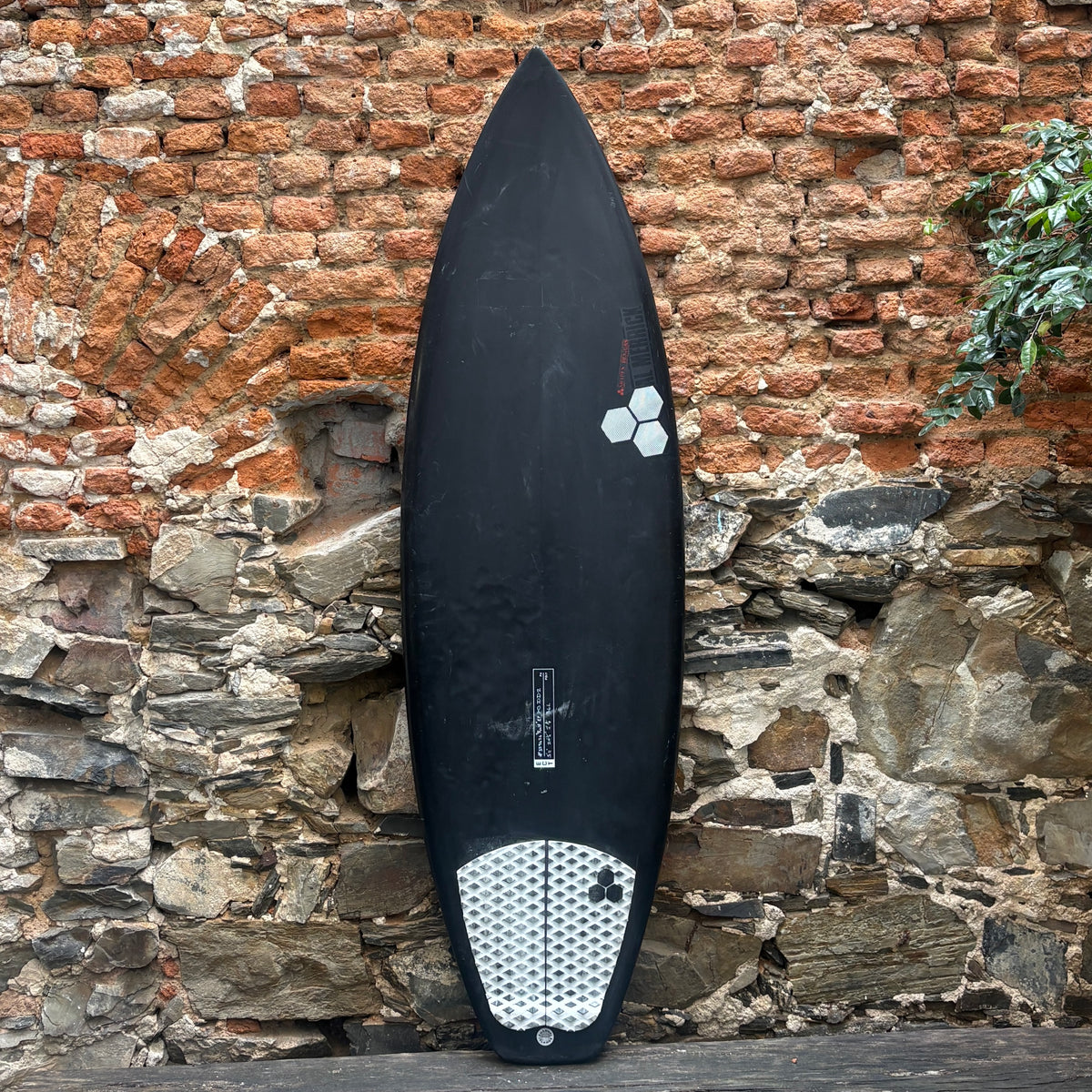 A black surfboard with a white hexagonal logo and a white grip tape tail, leaning against a rustic brick and stone wall.