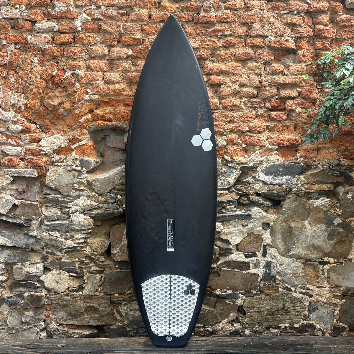 A black surfboard with a white and gray patterned tail pad, leaning against a rustic brick and stone wall.