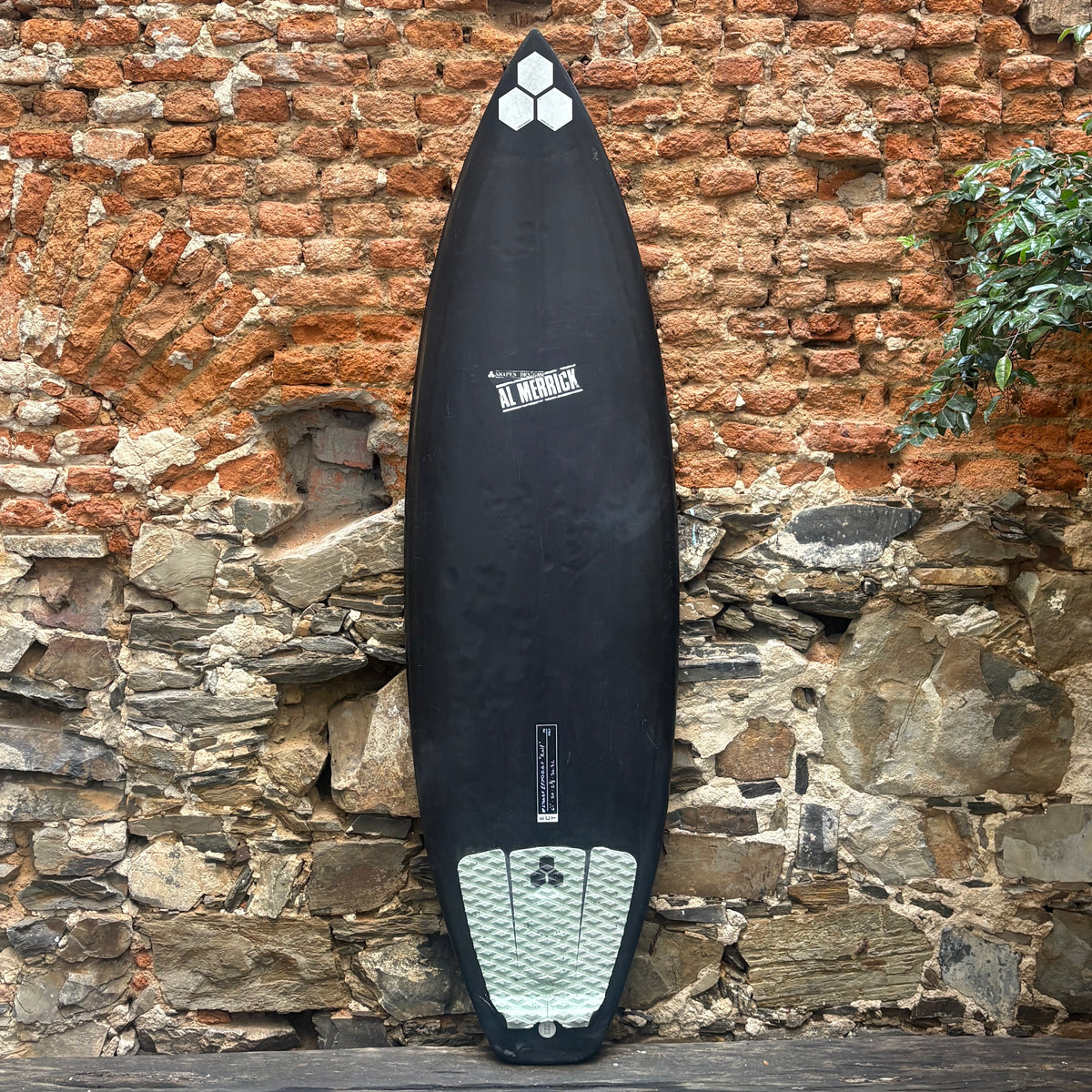 A black surfboard with white graphics and a white grip pad, leaning against a rustic stone and brick wall.