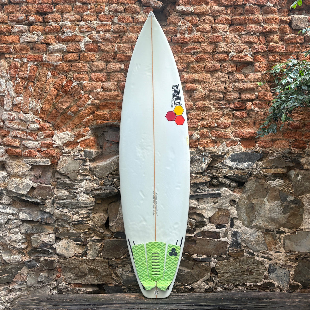 A white surfboard with a green traction pad and colorful logo decal, leaning against a rustic brick and stone wall.