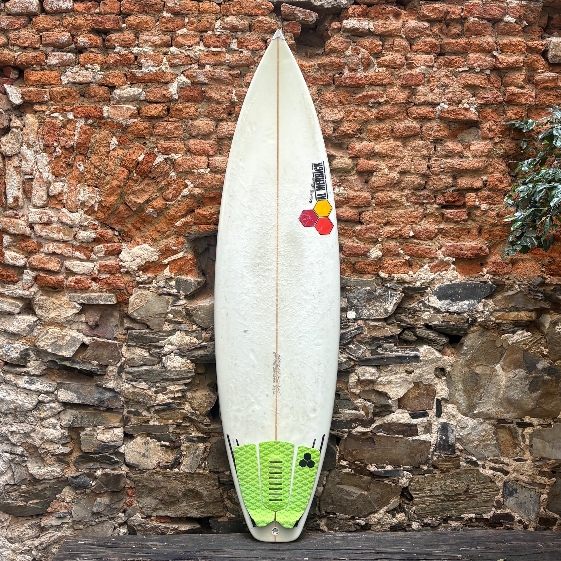 A white surfboard with a green and yellow tail pad and a black, white, and red logo decal, leaning against a rustic brick and stone wall.