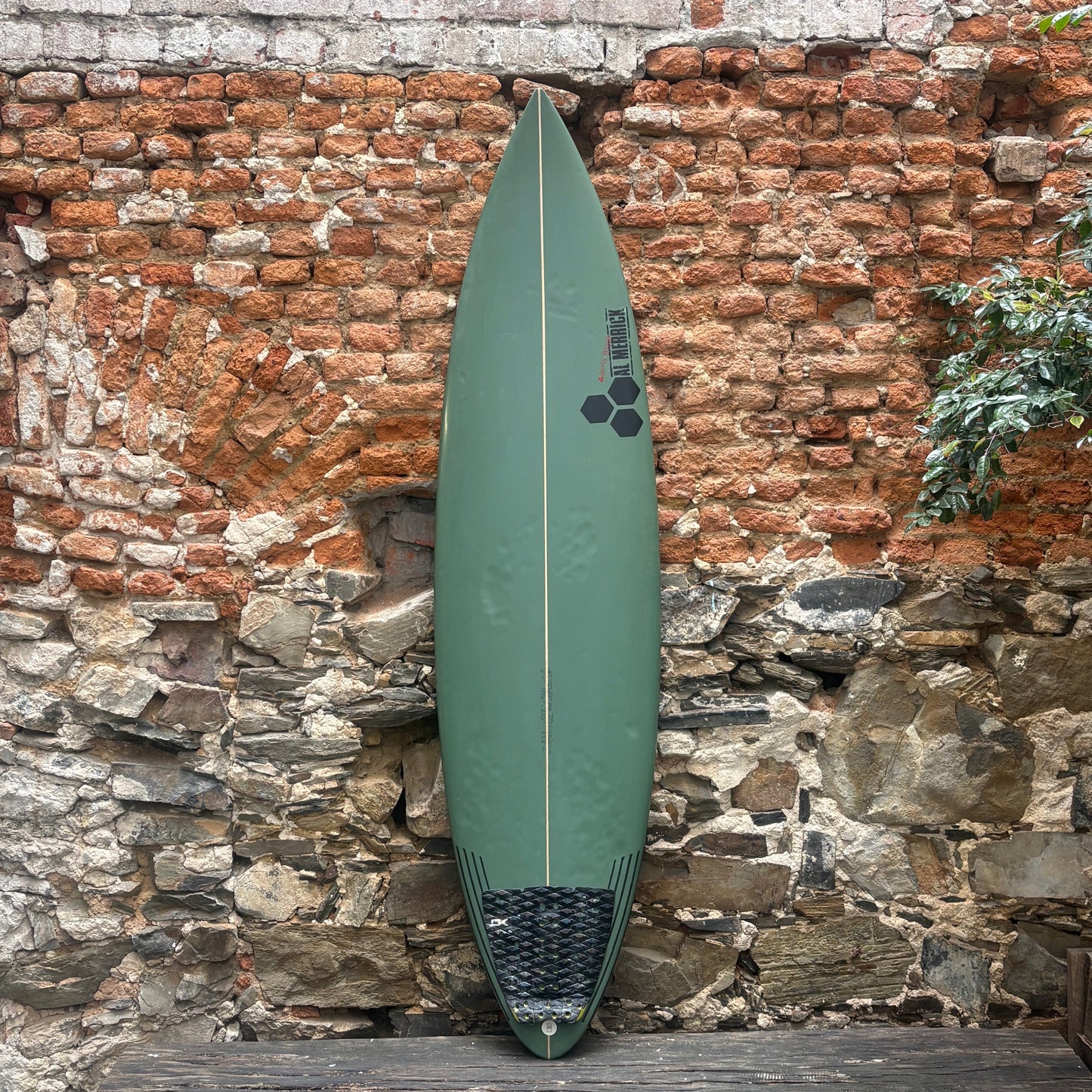 An olive green surfboard with a black hexagonal logo and a black grip pad at the tail, leaning against a rustic brick and stone wall.