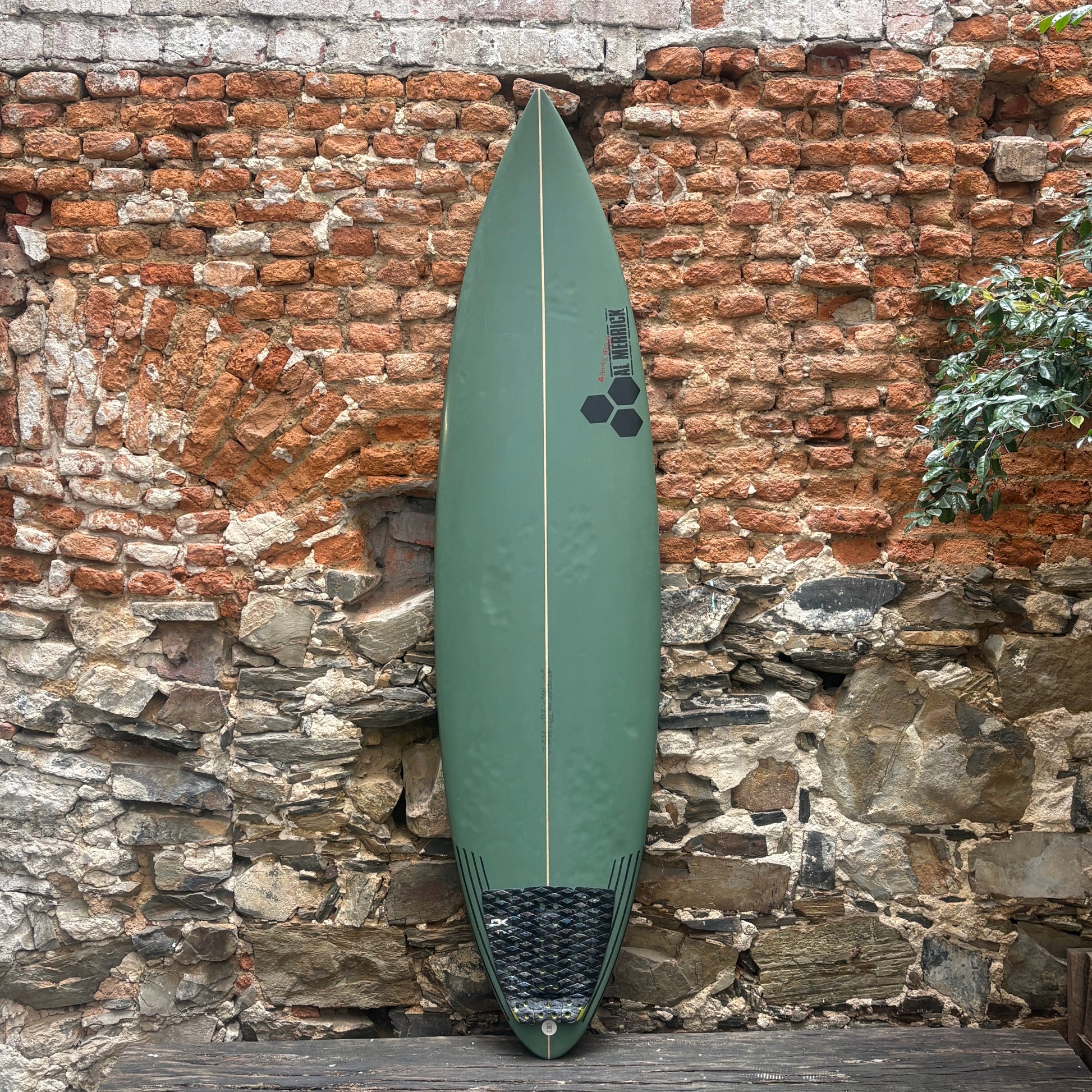 An olive green surfboard with a black hexagonal logo and a black grip pad at the tail, leaning against a rustic brick and stone wall.