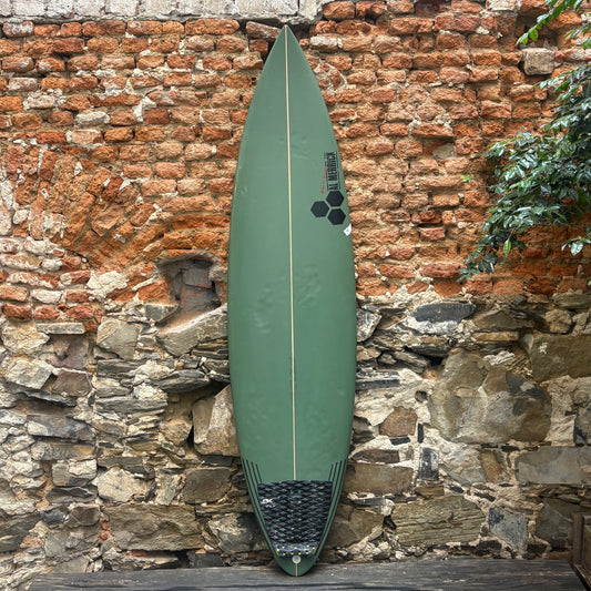 An olive green surfboard with a black hexagonal logo and a black grip pad at the tail, leaning against a rustic stone and brick wall.