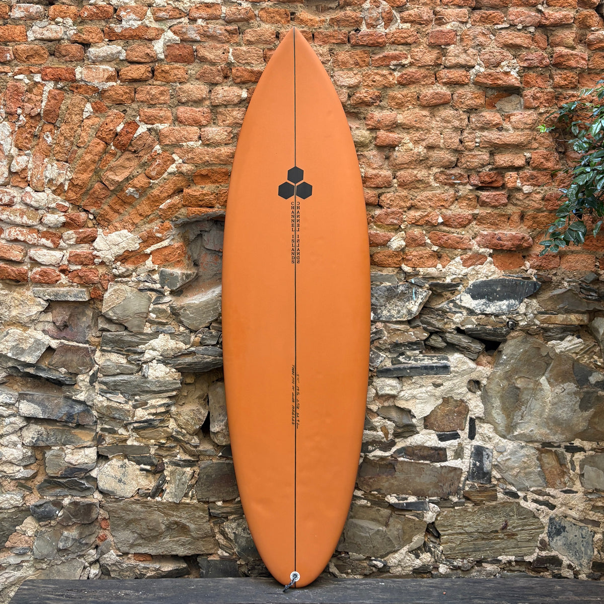 An orange surfboard with a black graphic design and text, leaning vertically against a rustic brick and stone wall.