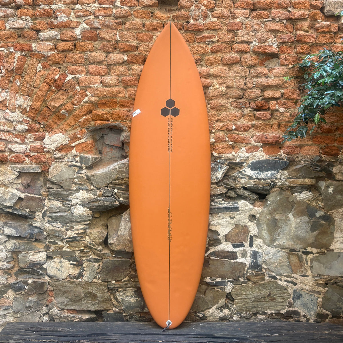 An orange surfboard with a black central graphic and text, leaning vertically against a rustic brick and stone wall.
