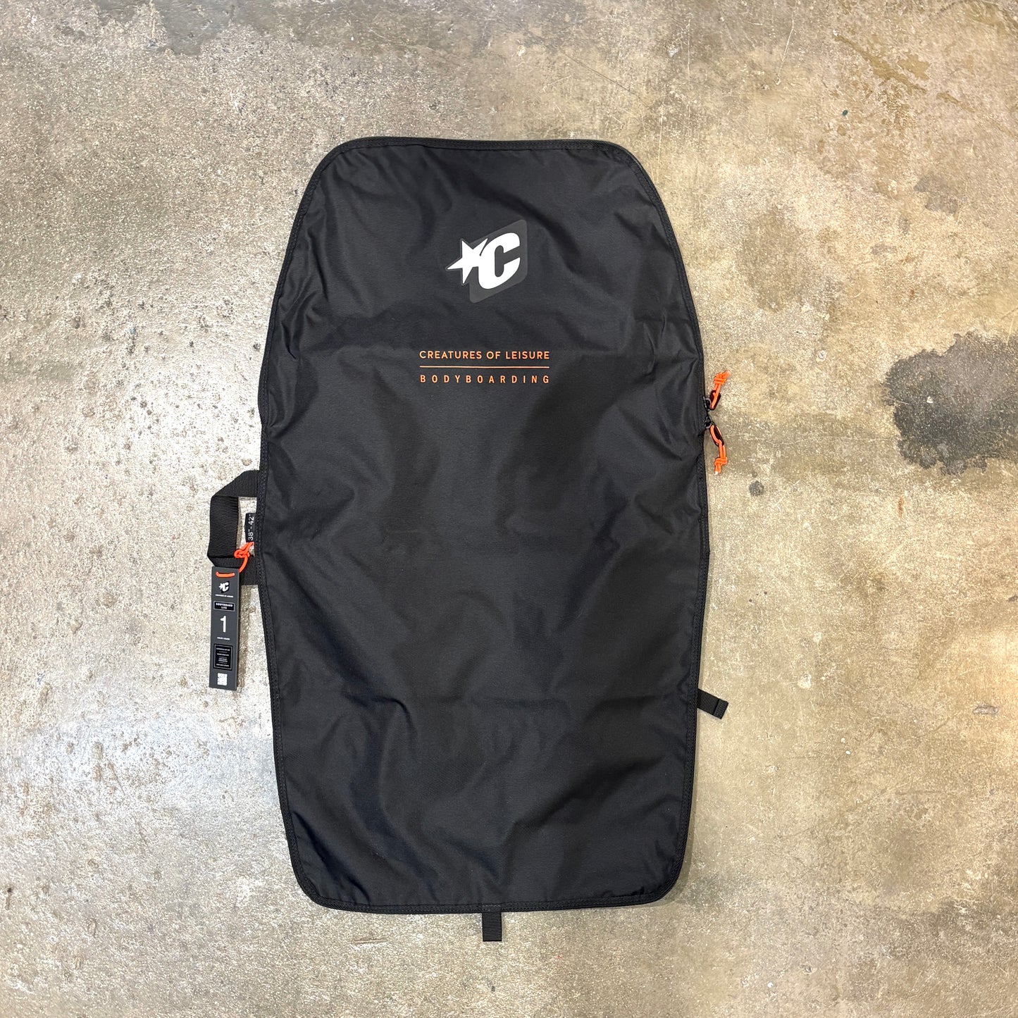 A black bodyboard bag laid flat on a concrete floor, featuring a white 'C' logo and 'Creatures of Leisure' branding with orange text accents.