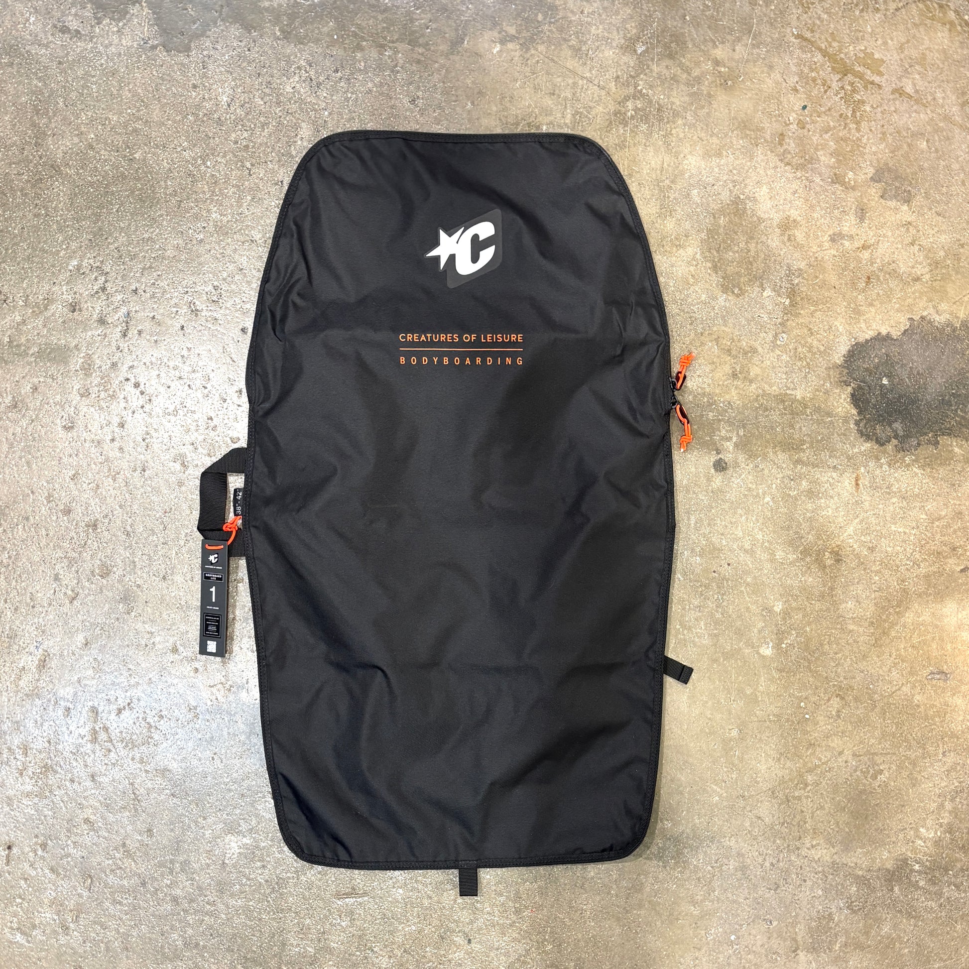 A black bodyboard bag laid flat on a concrete floor, featuring a white 'C' logo and 'Creatures of Leisure' branding with orange text accents.
