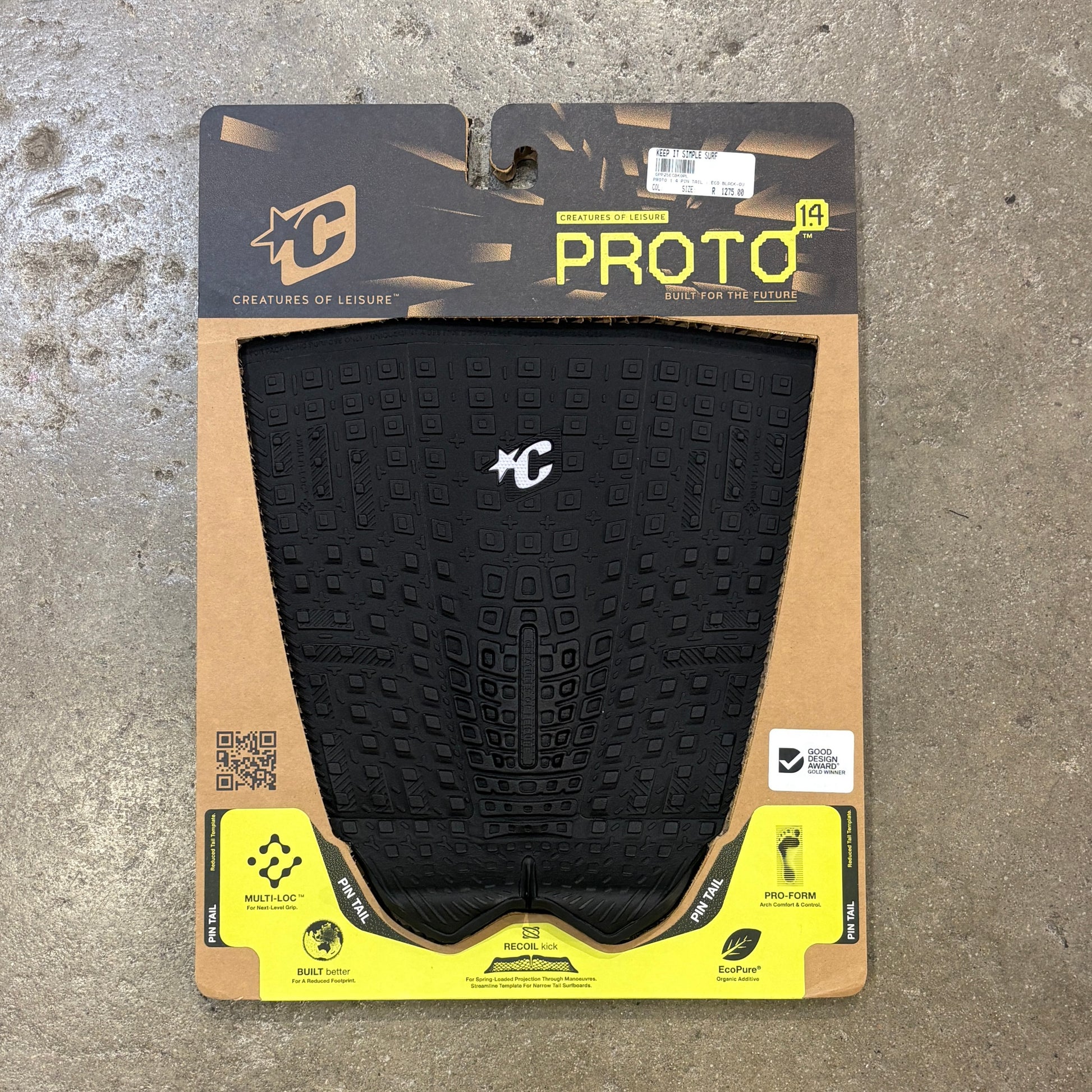 A black Proto 1.5 surfboard deck grip  package by Creatures of Leisure, displayed on a textured gray concrete floor. The grip tape is black with a textured grid pattern and a white 'C' logo, mounted on a cardboard backing with a black and yellow header.