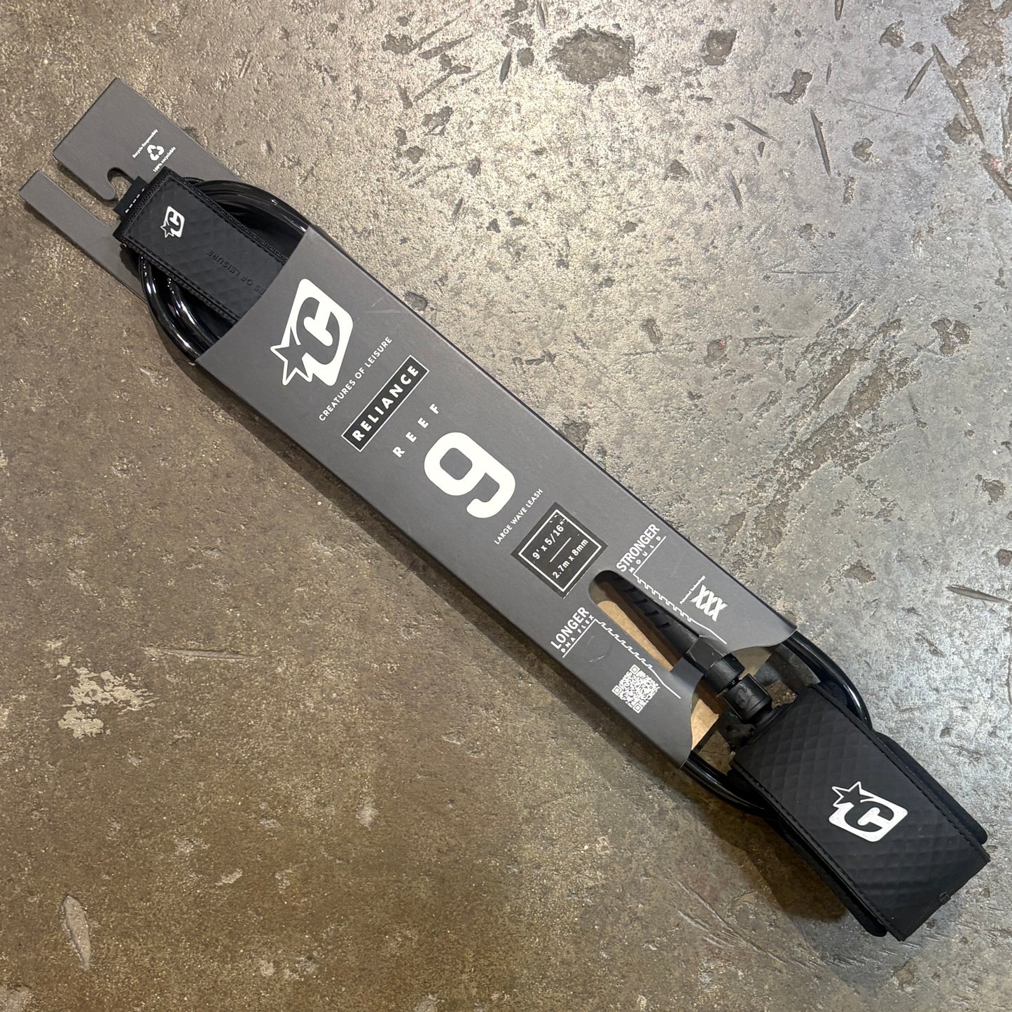 A black and gray surfboard leash packaged on a cardboard hanger, photographed diagonally on a textured concrete floor. The leash features a carbon-fiber patterned strap and a black buckle.