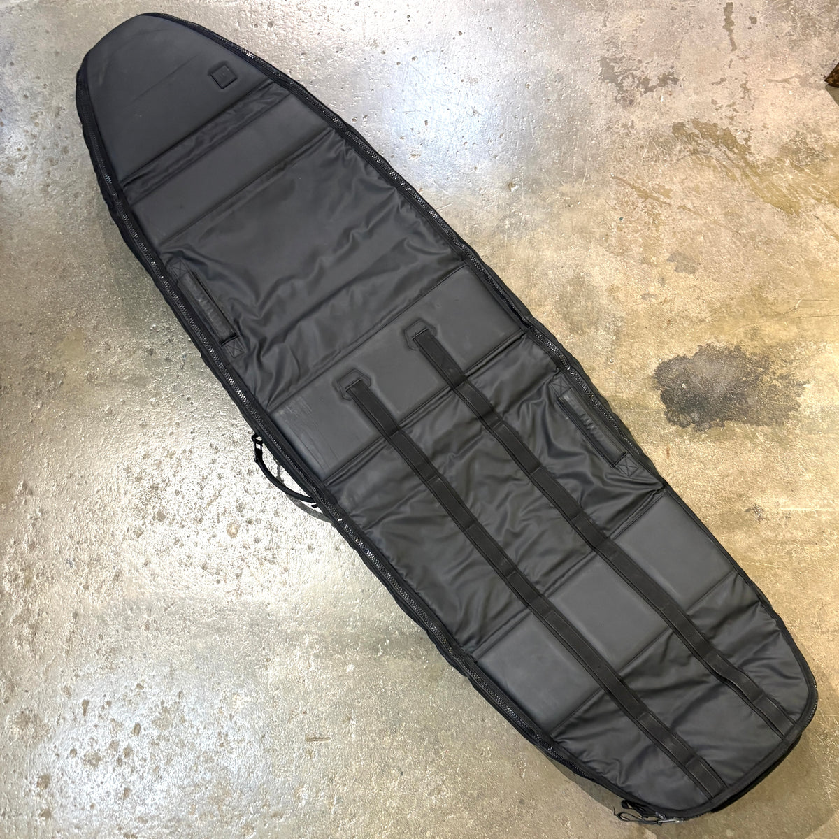 A black padded surfboard bag laid diagonally on a concrete floor.
