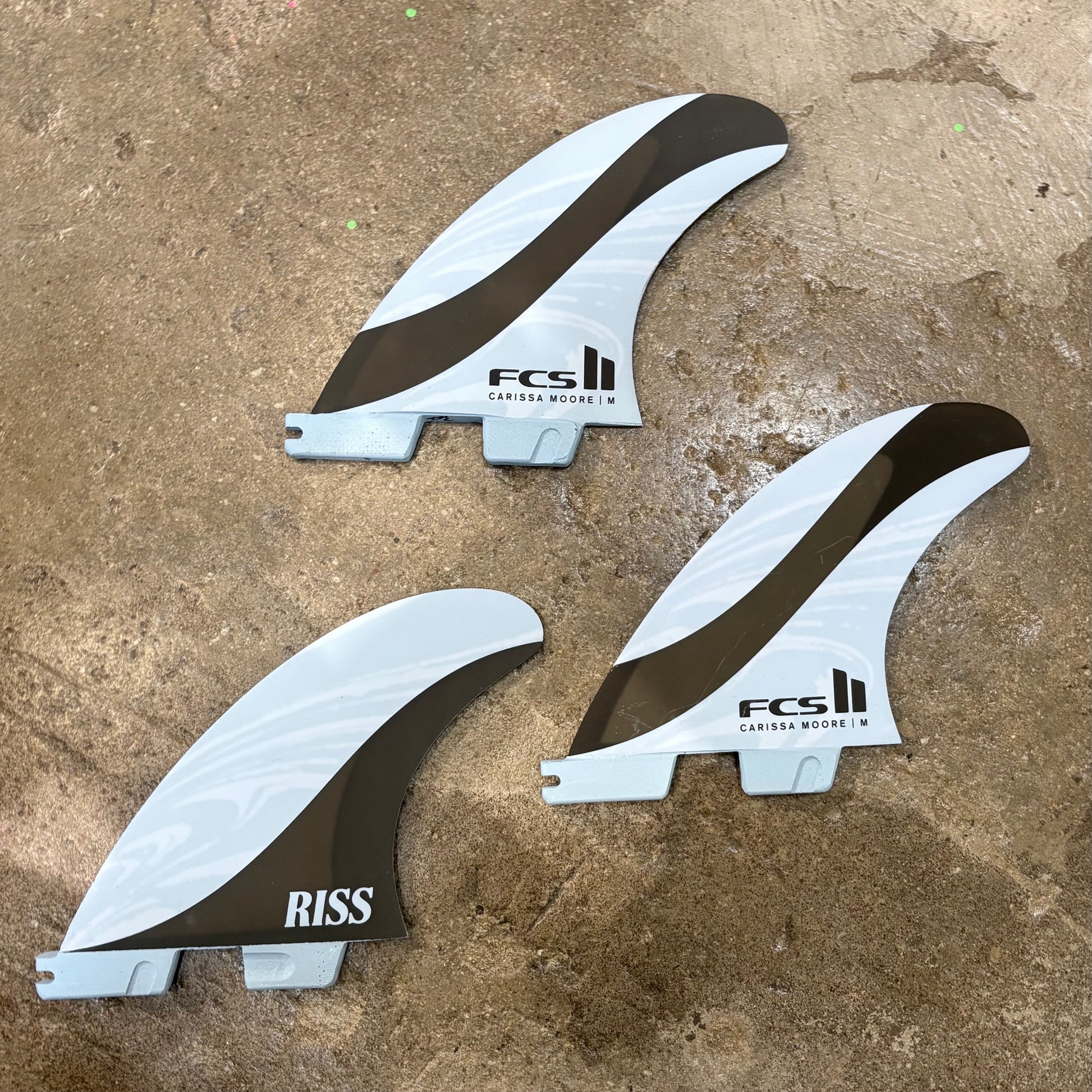 FCS II Carrisa Moore Signature Tri Fin Set - Blue and Black - Full thruster set laid on concrete floor