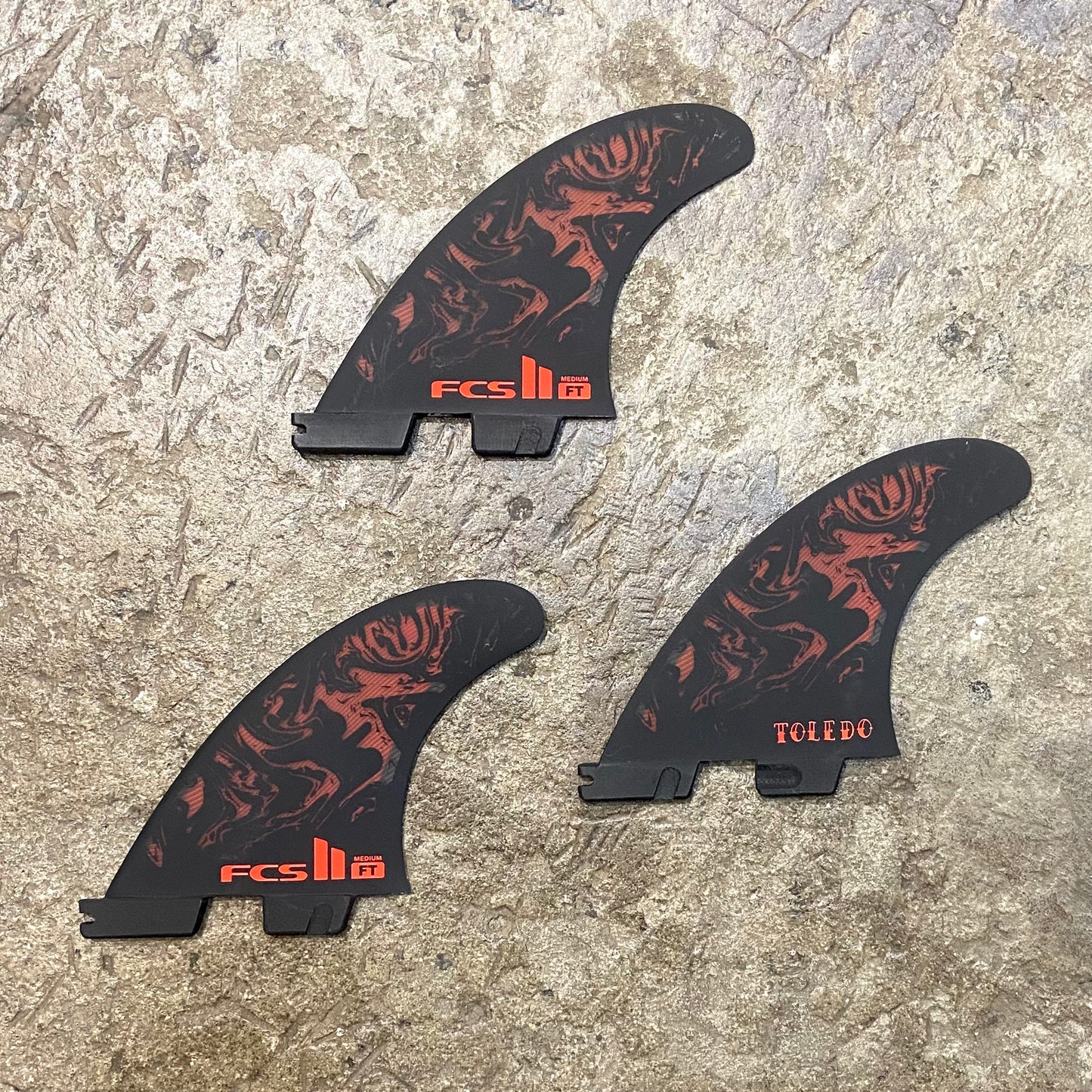 View of FCS II Filipe Toledo Tri Fins L in a red and black colour way laid out on a concrete floor