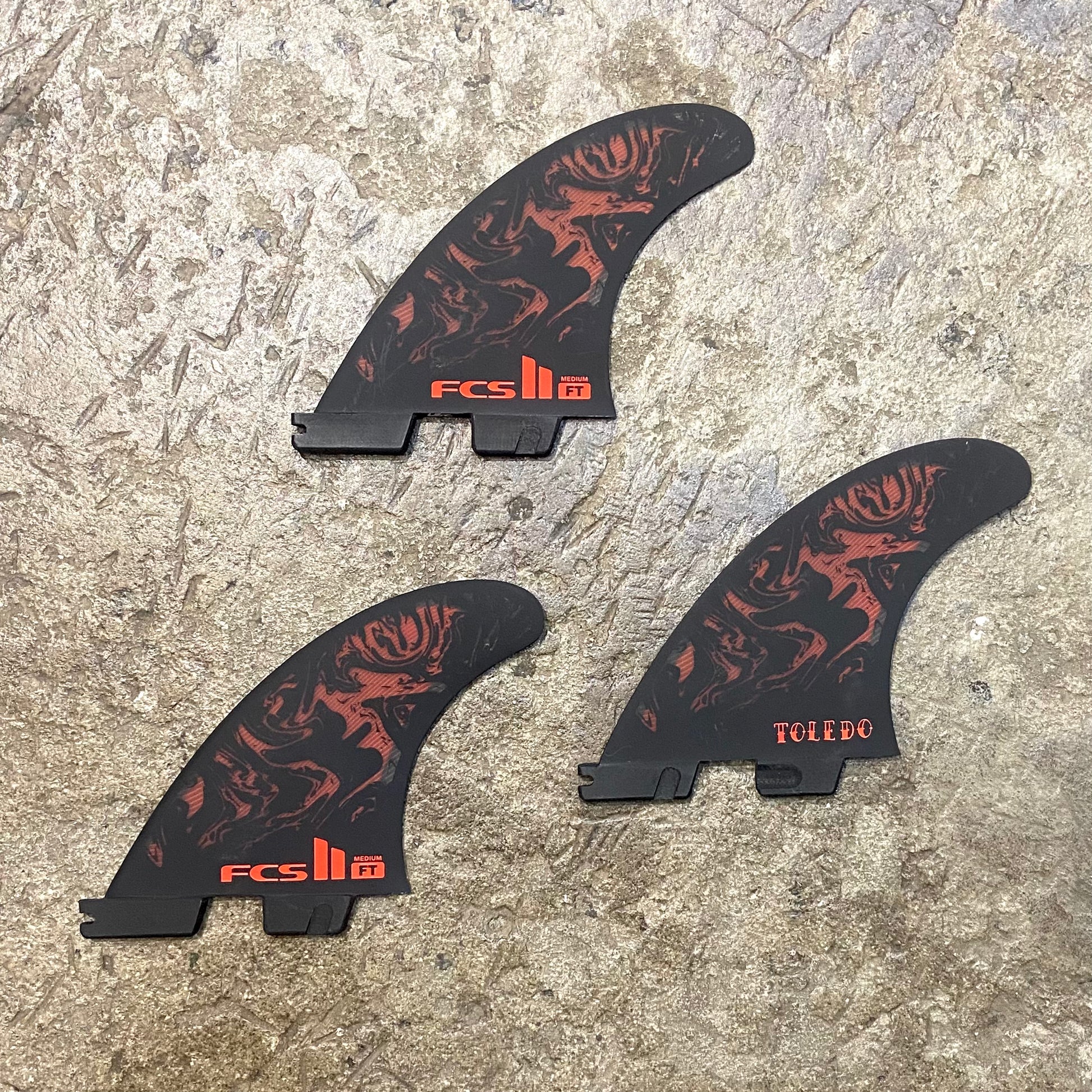 View of FCS II Filipe Toledo Tri Fins L in a red and black colour way laid out on a concrete floor