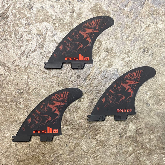 View of FCS II Filipe Toledo Tri Fins L in a red and black colour way laid out on a concrete floor