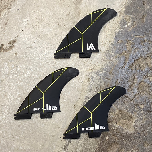 A set of medium FCS II Kolohe Andino PC Tri fin in a black colour way with green details on a concrete floor