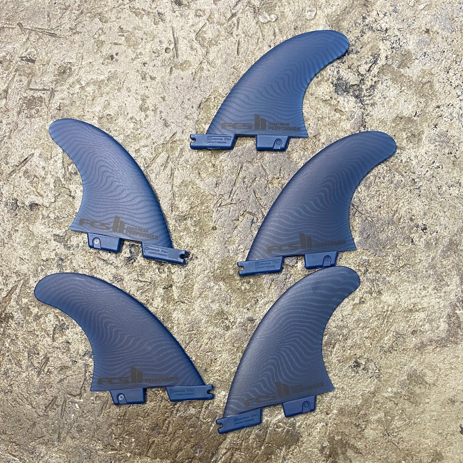View of the FCS II Performer Neo Eco Tri-Quad Fin Set M, all five fins in a blue colourway laid out on a concrete floor