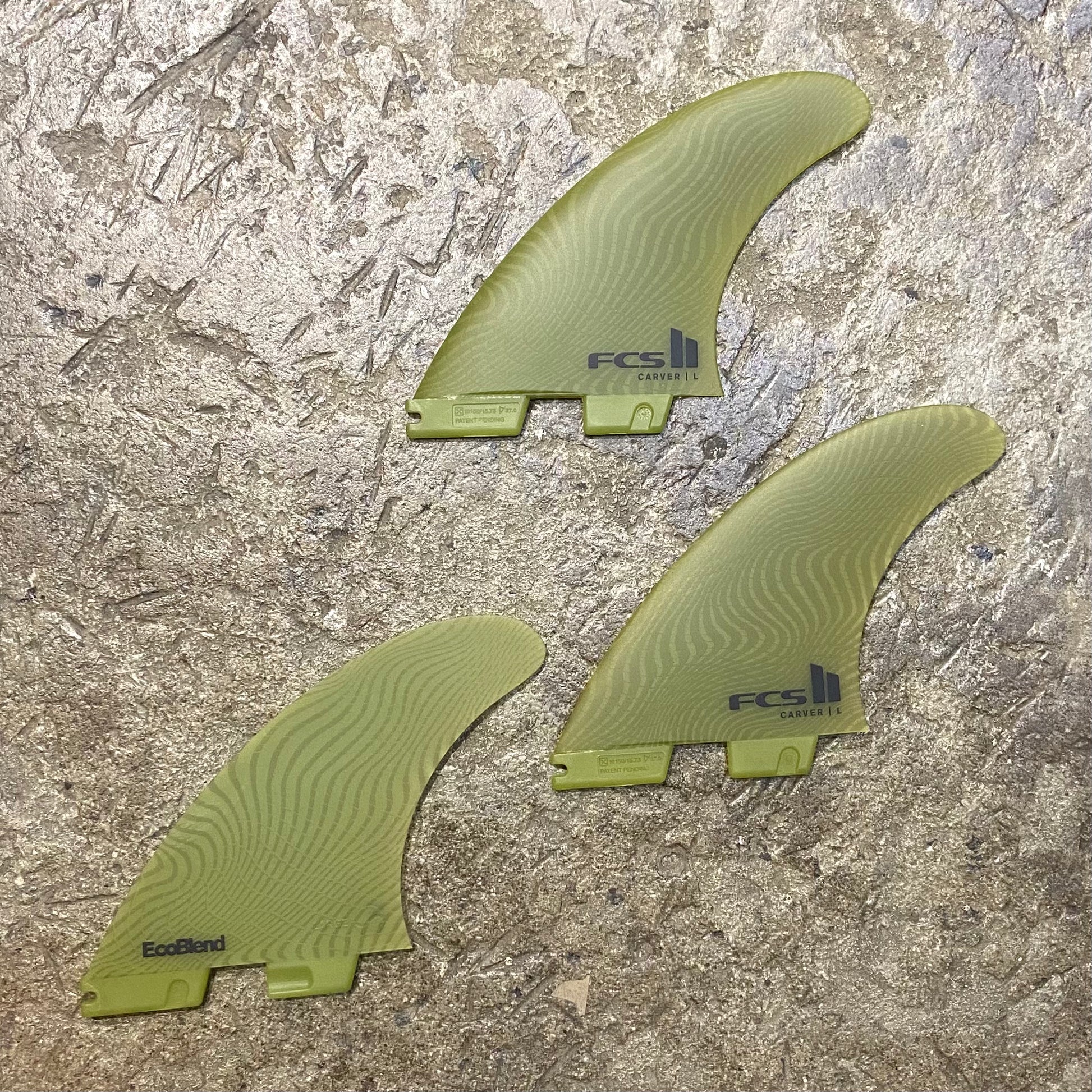 A set of the FCS ll Carver Neo Glass Eco Tri Fins in the green colour way laid out on a concrete floor