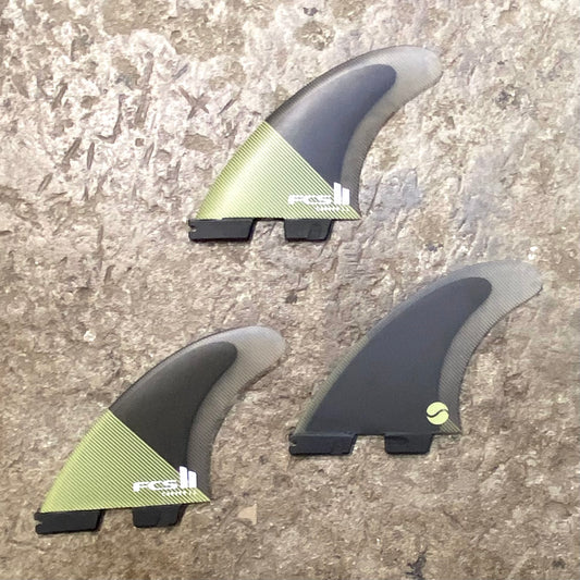 A large set of FCS ll Essential Series Carver PC Tri Fins in a black and green colour way laid on a concrete surface