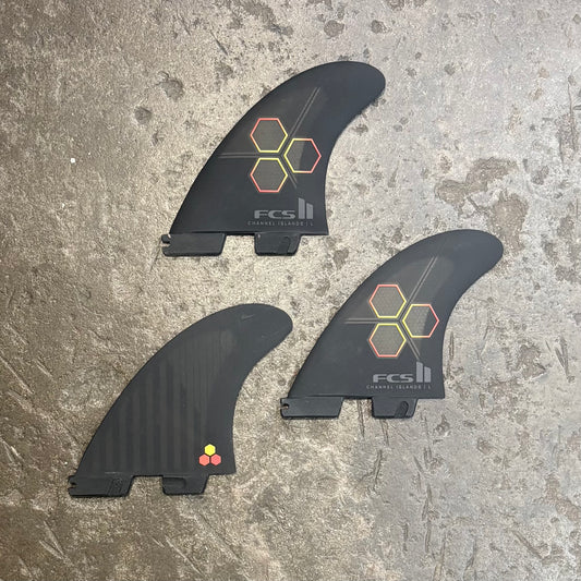 Three black surfboard fins with FCS II branding on a textured surface