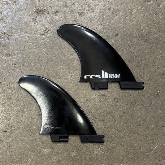 Two black surfboard tail fins, one branded FCS Quad Rear Carver and the other FCS Speed, placed on a textured concrete floor.