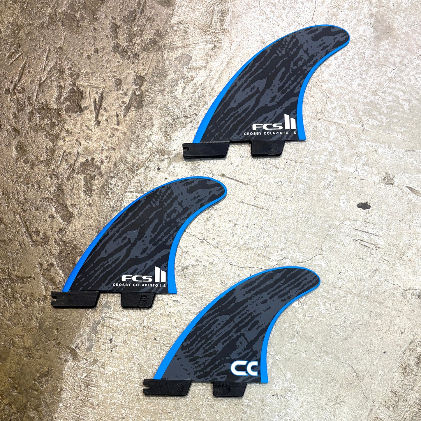 Three FCS II surfboard fins with a black and blue wave pattern, displayed on a textured concrete surface.