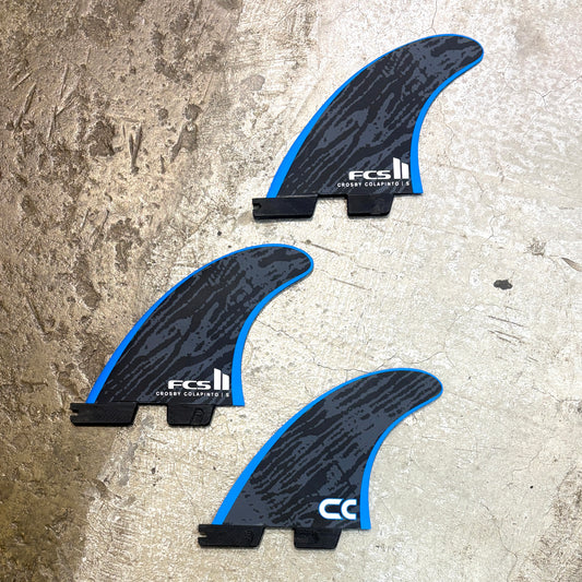 Three FCS II surfboard fins with a black and blue wave pattern, displayed on a textured concrete surface.