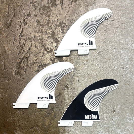 Three FCS surfboard fins arranged on a textured concrete surface, including two white fins and one black fin with the brand name MEDINA visible.