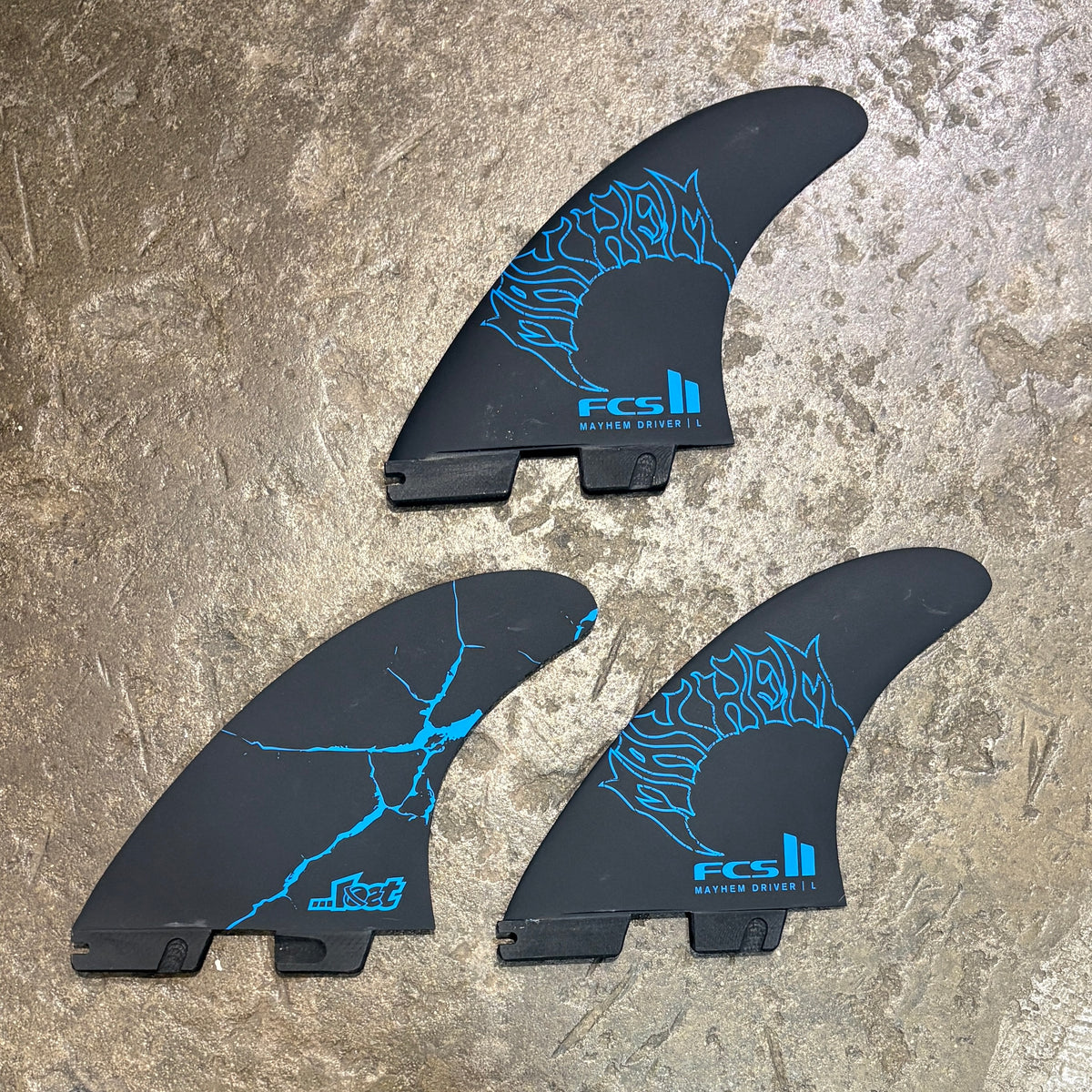 Three black surfboard fins with bright blue graphic designs and FCS branding, arranged on a textured stone surface.