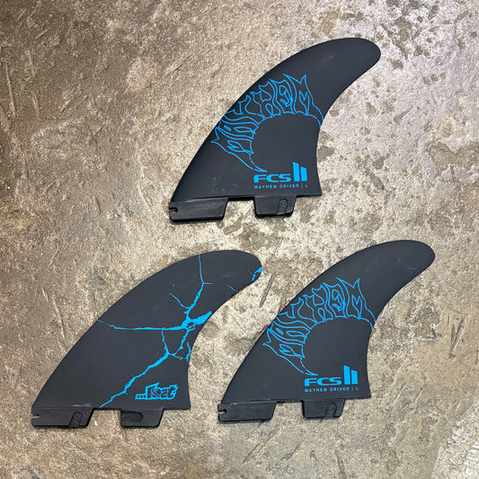 Three black surfboard fins with bright blue graphic designs and FCS branding, arranged on a textured stone surface.