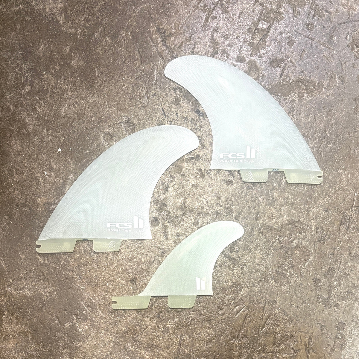 Three translucent white surfboard fins arranged on a textured brown stone surface. The fins show a wood grain pattern and the brand logo 'FCS' is visible on two of the fins.