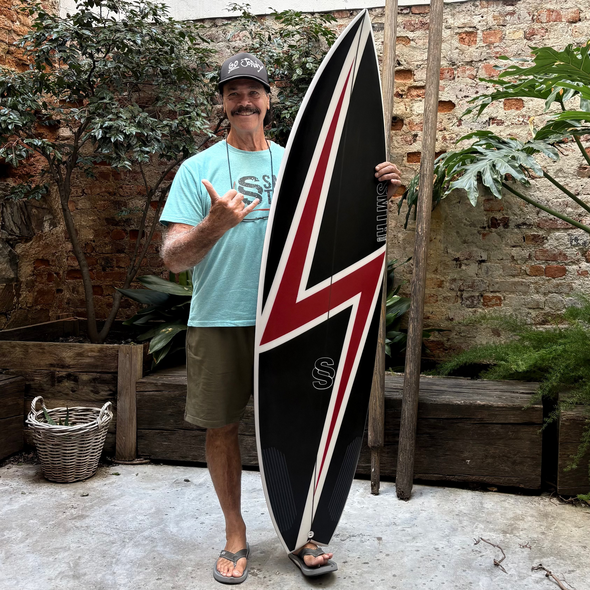 Man holding a black and red surfboard against a brick wall.