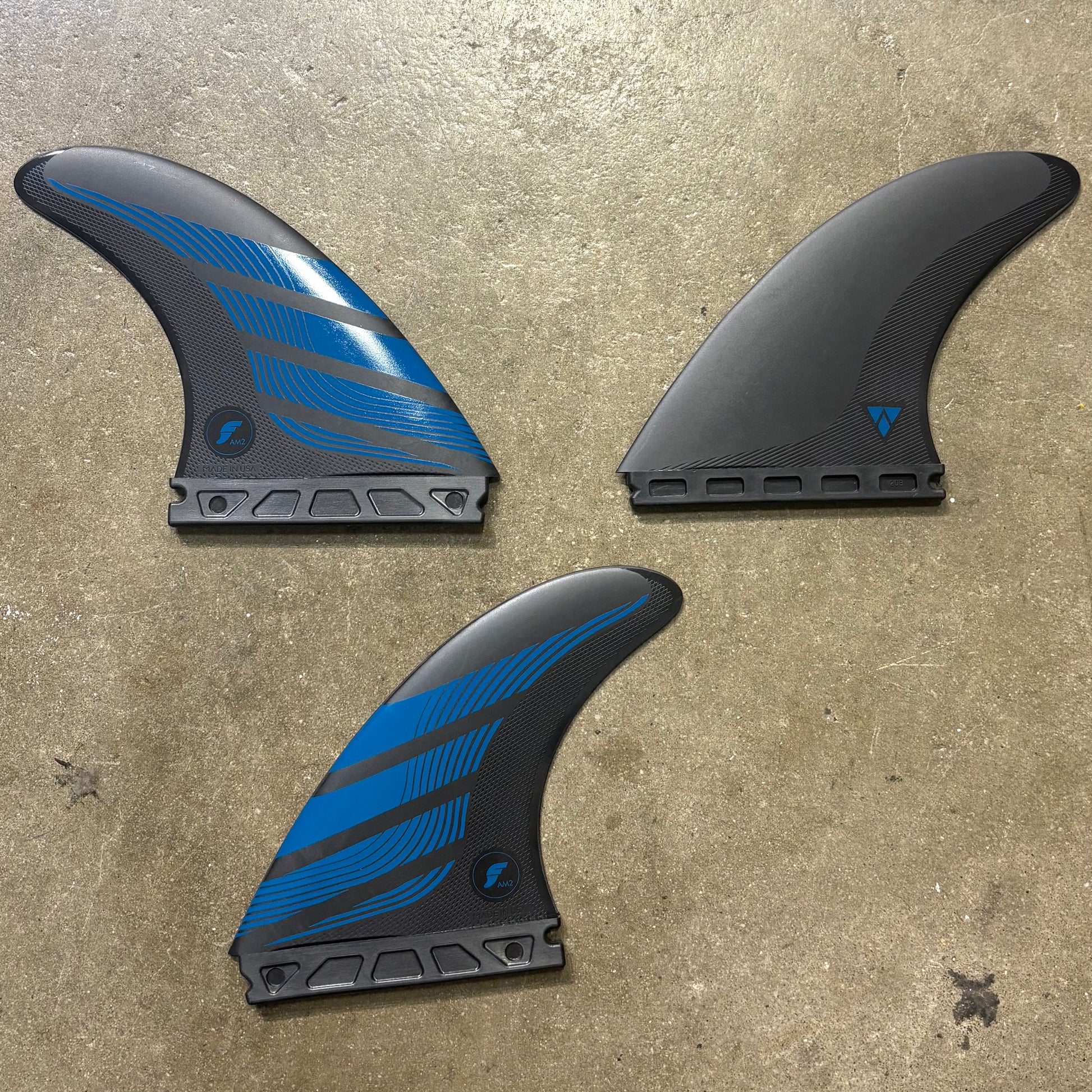 A set of Futures Alpha AM2 Surf fins in a black and blue colour way laid out on a textured concrete surface