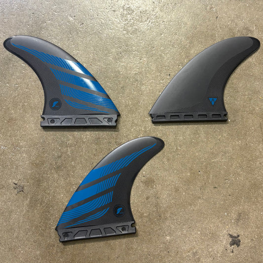 A set of Futures Alpha AM2 Surf fins in a black and blue colour way laid out on a textured concrete surface