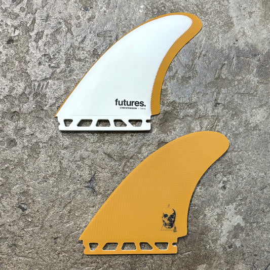 A pair of futures The Christenson Twin fins, one with white and the other with a yellow color scheme, displayed on a textured surface.