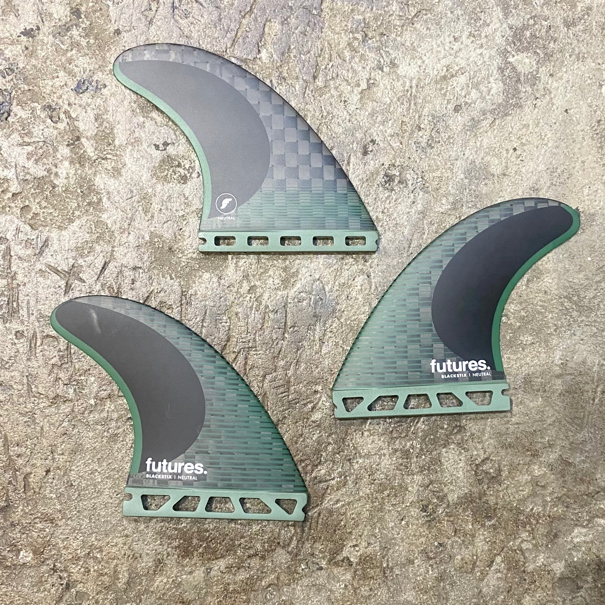 A set of Futures F8 Blackstix black and green surfboard fins with the futures brand logo on them, displayed on a textured grey surface.
