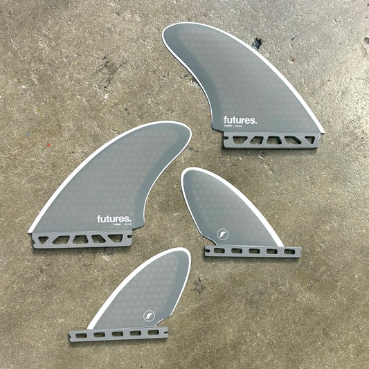 Four grey surfboard fins from the Flow Quad Set with 'futures' branding on a concrete surface.