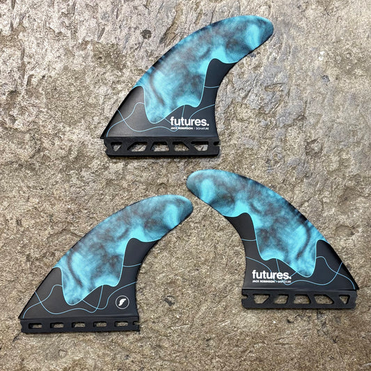 A set of Jack Robinson signature Futures surfboard fins with a honeycomb design, predominantly black with blue accents, laid on a concrete surface.