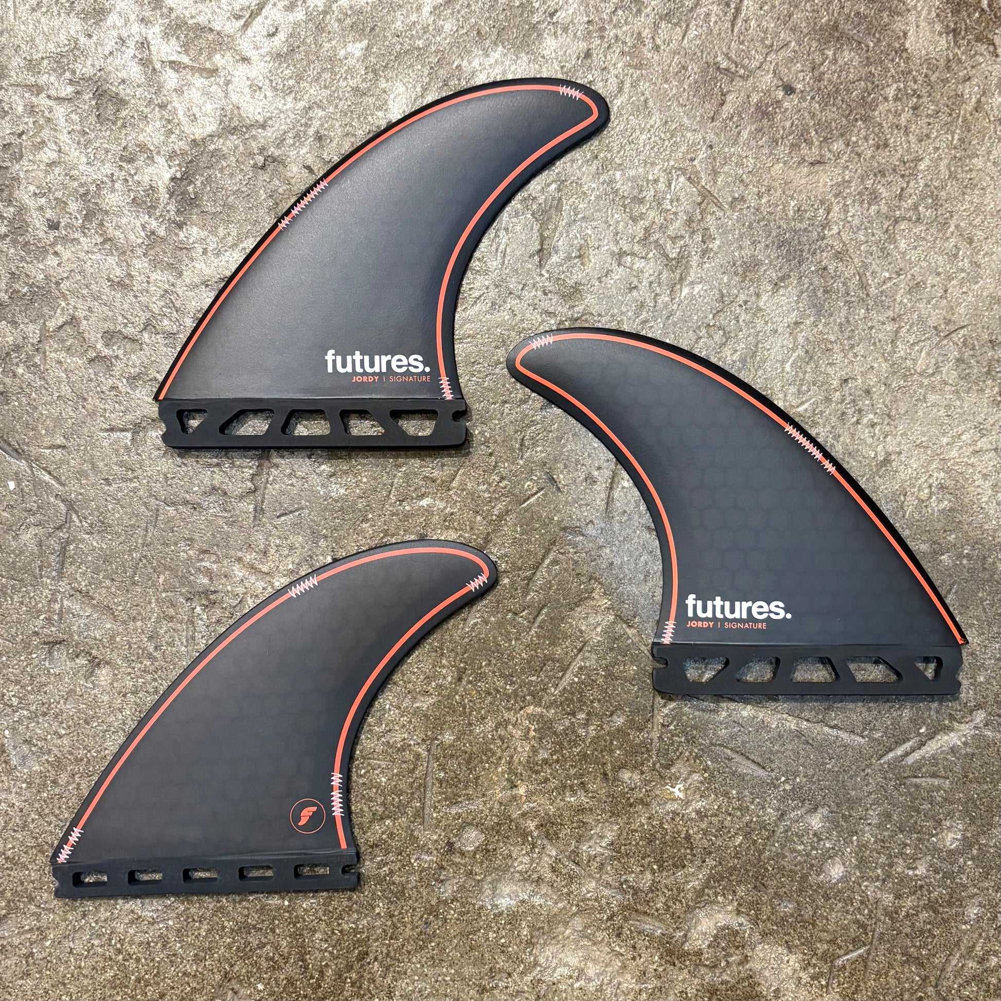 Three black Futures Jordy Signature surf fins with red accents on a concrete surface.