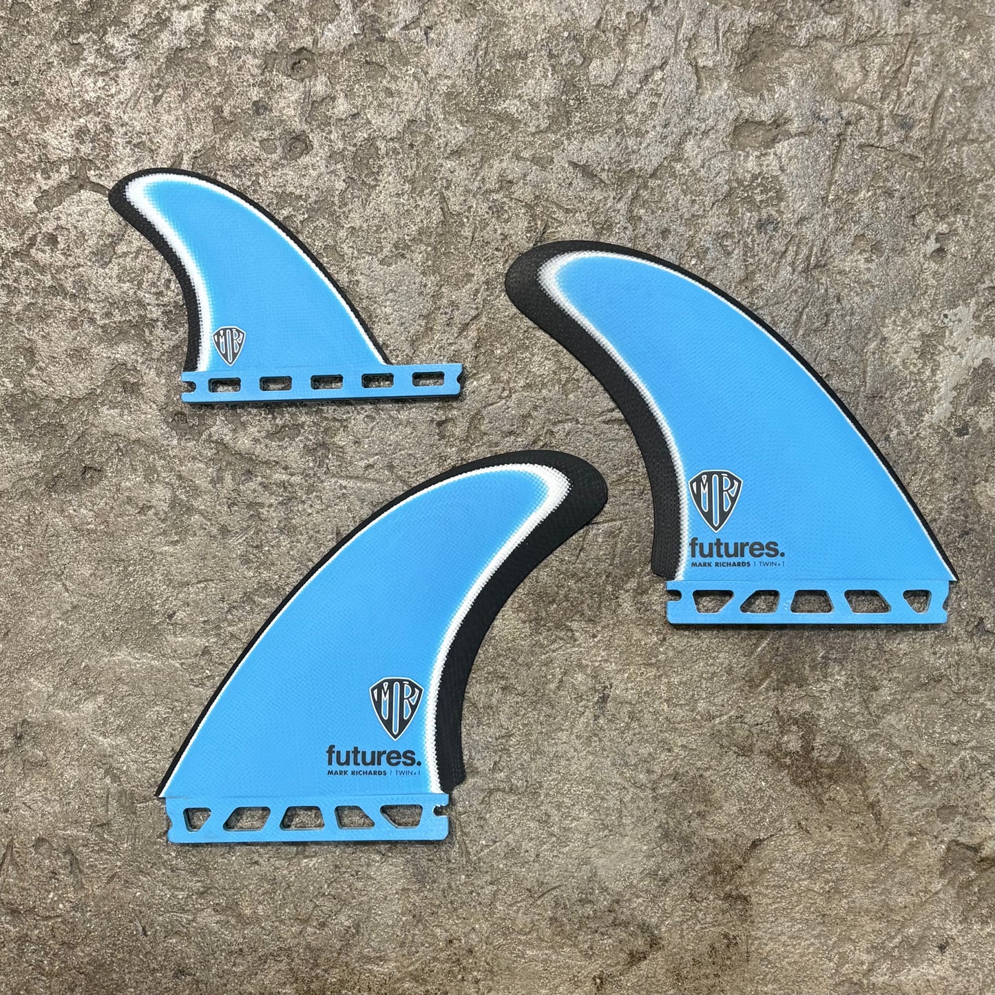 A set of Three blue Futures Mark Richards Twin+1 surfboard fins with on a concrete surface