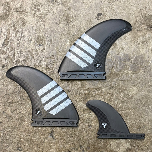 A set of black Futures Rasta Alpha Twin+1 surfboard fins with silver stripes on a textured grey background.