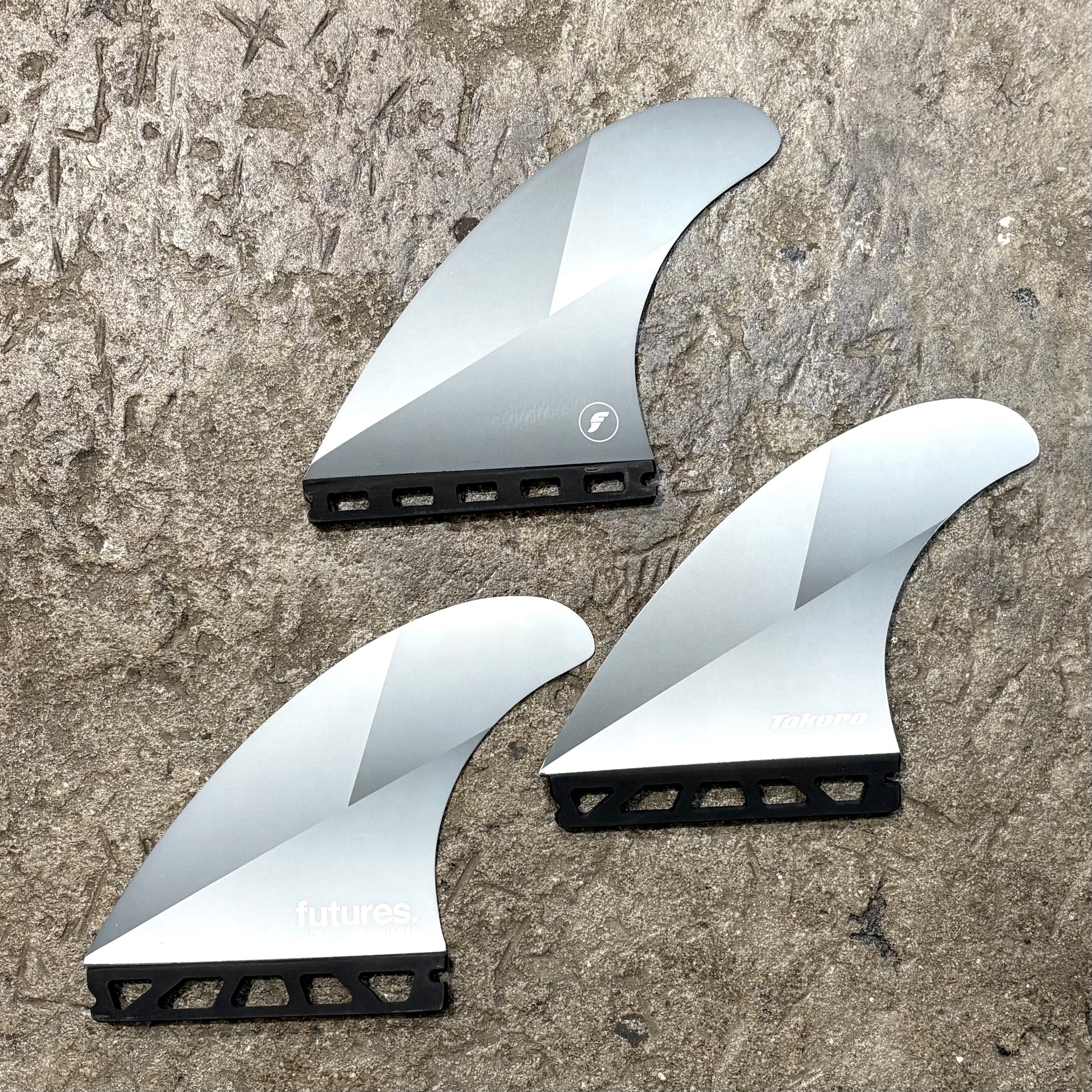 A set of Futures Tokoro silver surfboard fins with honeycomb construction, laid on a textured ground.