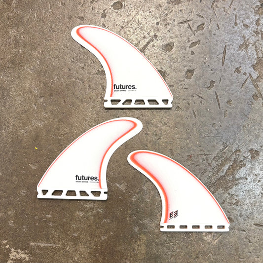 Three white Ethan Ewing surfboard fins with red accents on a textured surface, featuring the 'futures' brand.