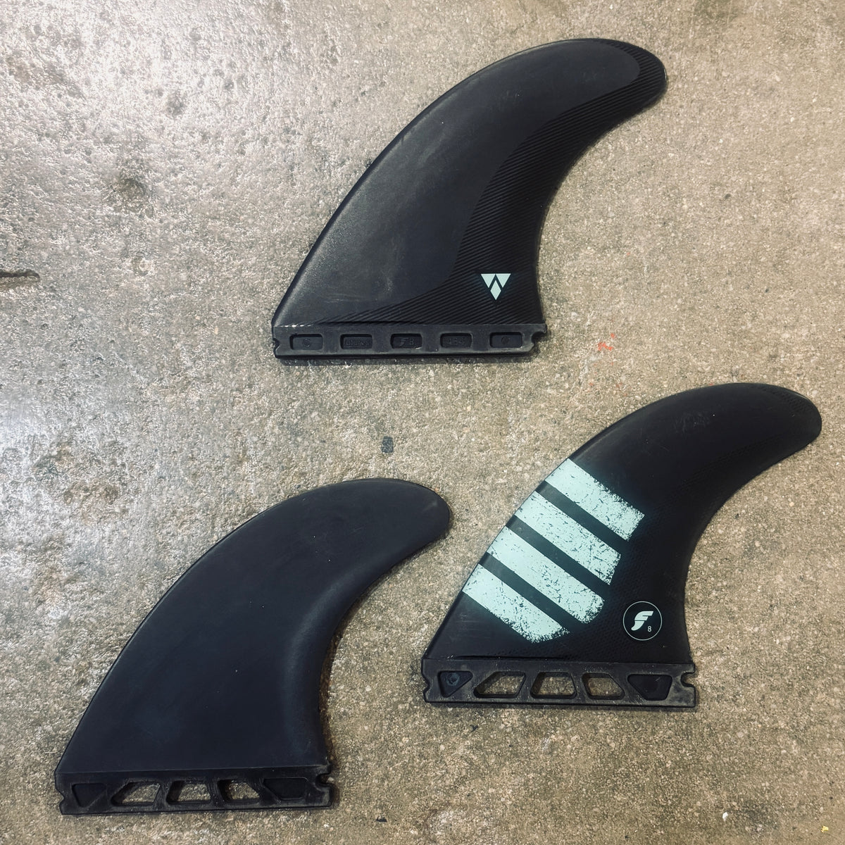 Three black carbon fiber surfboard fins arranged on a concrete floor. One fin features three light blue/white painted stripes and a small black logo, and another has a small white geometric logo.