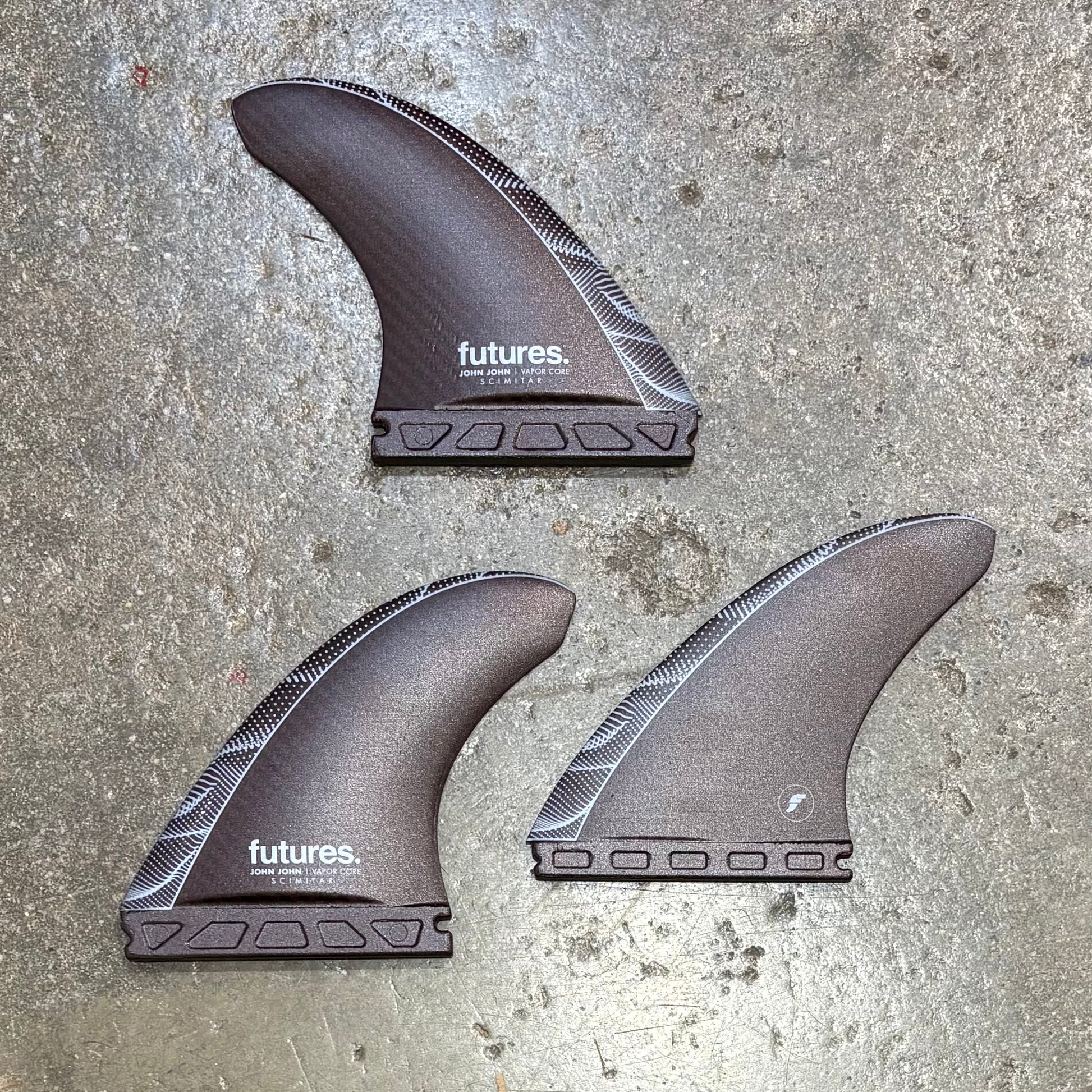 Three John John Florence surfboard fins with 'futures' branding on a concrete surface