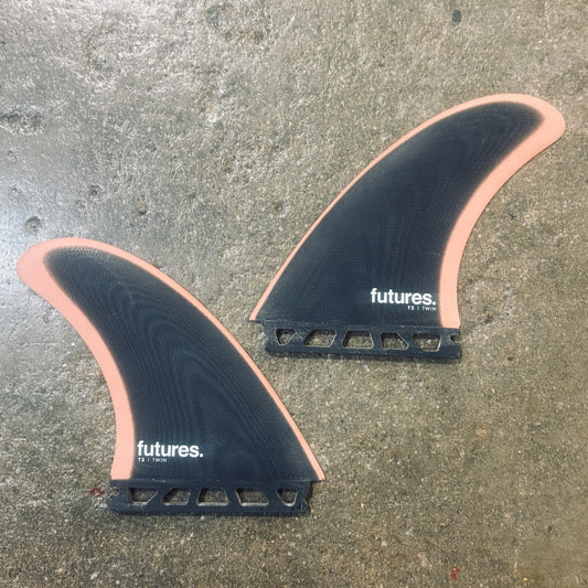 Two surfboard fins with a black carbon fiber body and a peach-pink edge, featuring the white 'futures.' logo, displayed on a textured concrete floor.