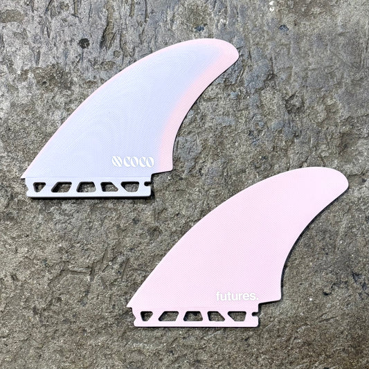 A set of Futures XO Coco Mod Keel surfboard fins with a light pink color and the branding 'XO COCO' visible on them, placed on a textured grey surface.