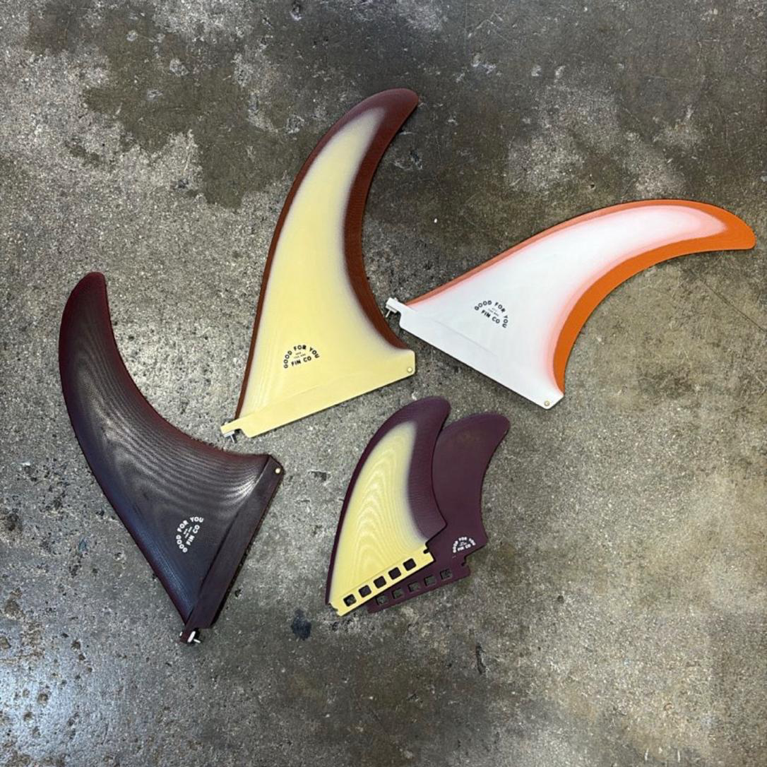 Set of four colorful surf fins on a concrete surface with 'Local Goodness' branding.