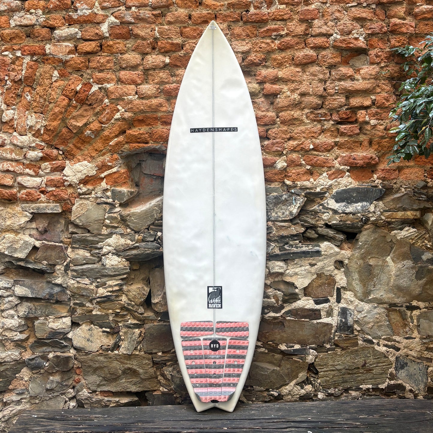 A white surfboard with a red and black striped tail patch, leaning against a rustic brick and stone wall.